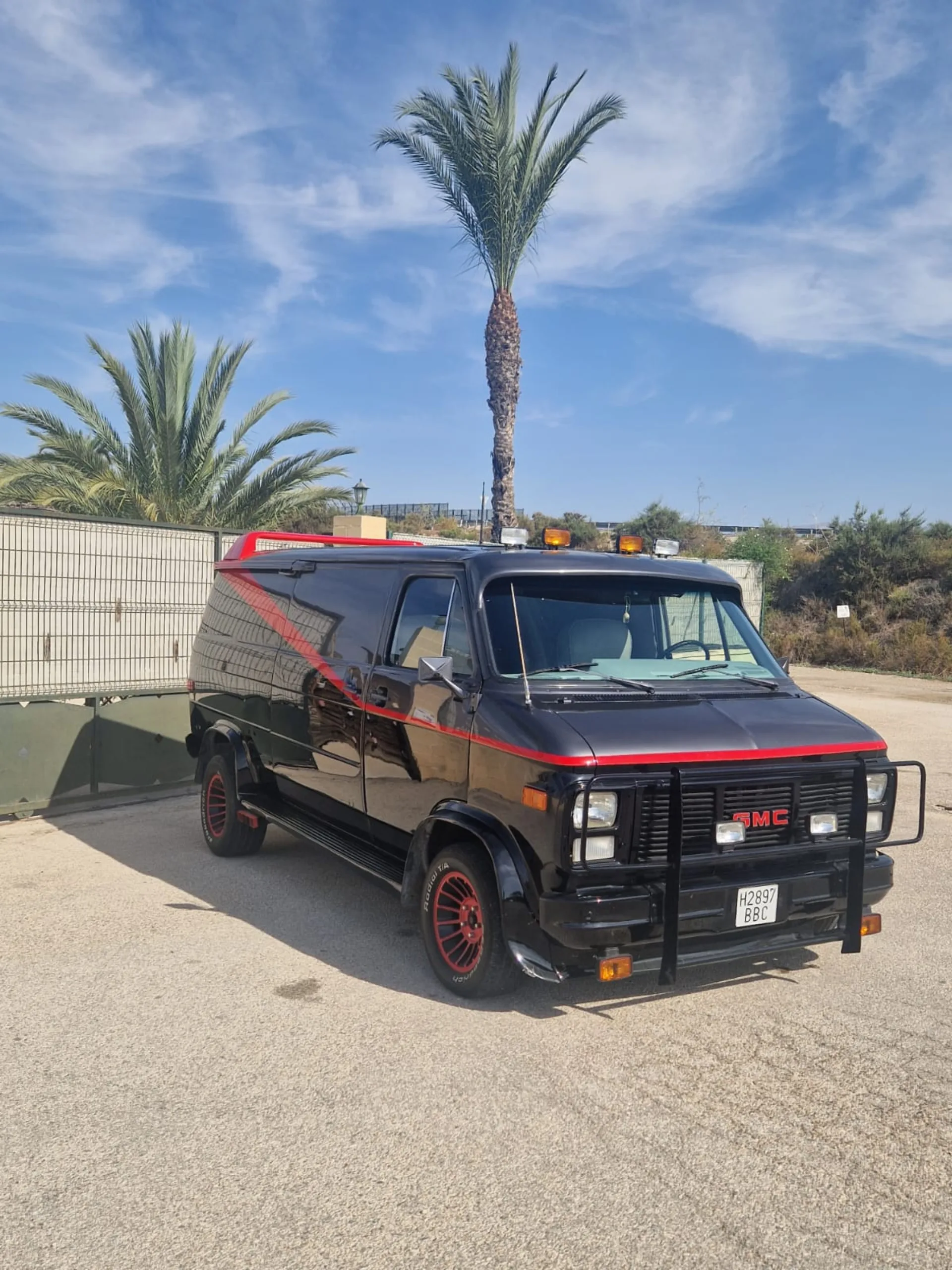 The black and red A-Team van, a GMC replica with bull bar and roof lights, under a palm tree with blue sky in Spain